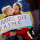 A child is holding a protest sign that says: “Putin, hands off Ukraine.” The child is sitting on their caregiver’s lap. Behind them is a Ukrainian flag. 
